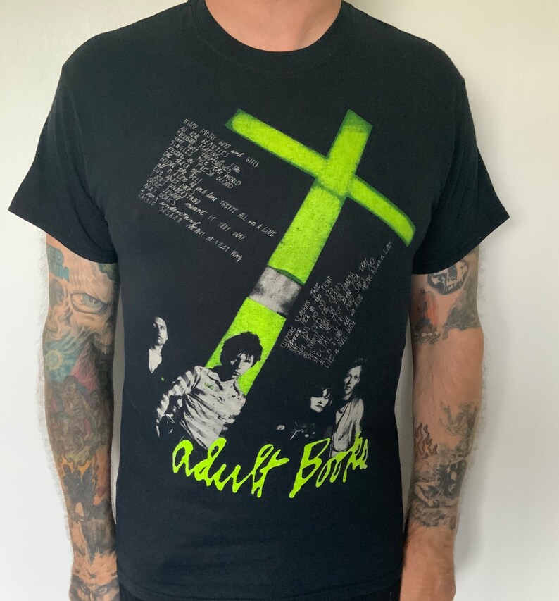 May include: Black t-shirt with a green cross graphic and the text "adult Books" printed on the front. The shirt features a band of lyrics printed on the side.
