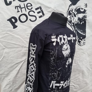 K&ouml;nnte beinhalten: Schwarzes Langarmshirt mit wei&szlig;em Druck. Die Vorderseite des Shirts zeigt eine Grafik einer Person mit dem Text "ライオター パーティション" in Japanisch. Der linke &Auml;rmel hat den Text "DESTROYERS" in wei&szlig; gedruckt.
