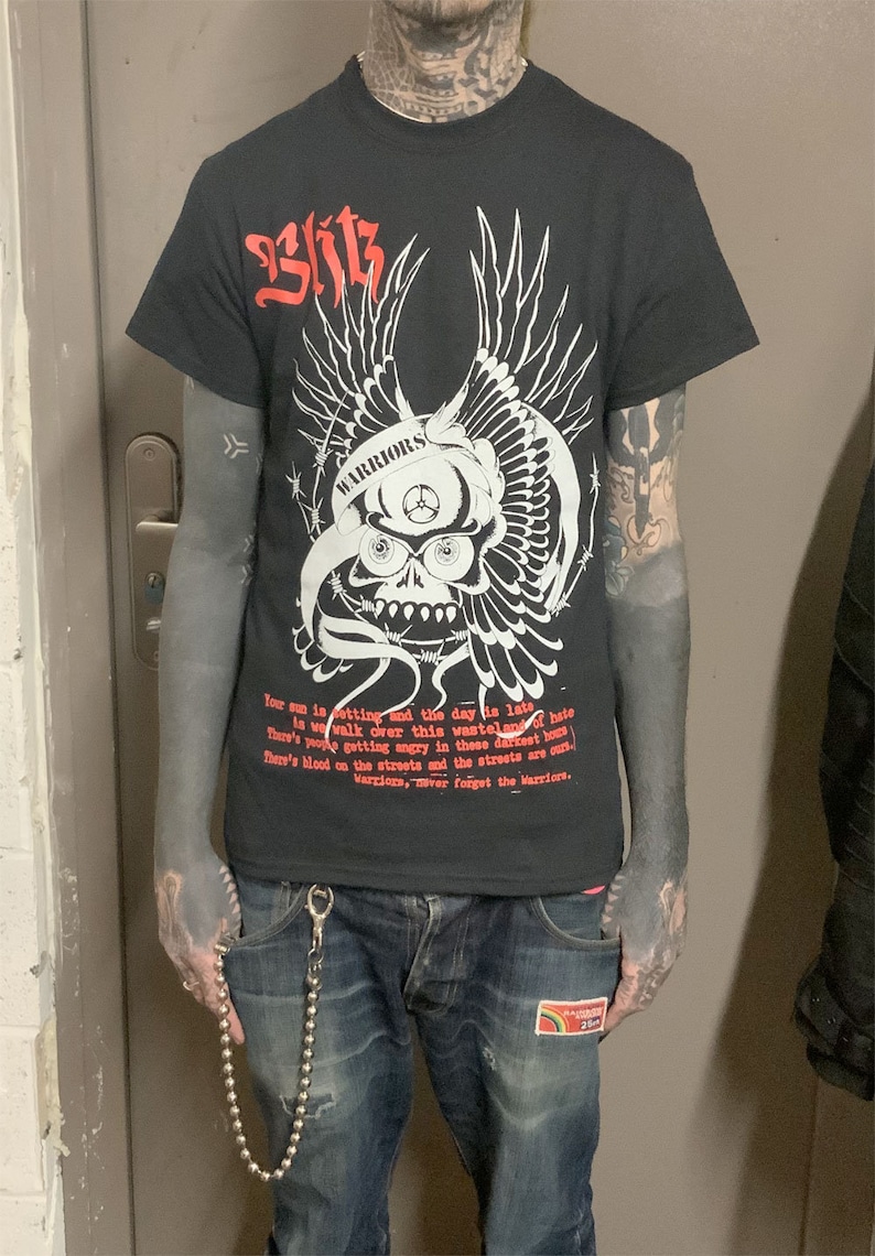 以下が含まれることがあります： 白い骸骨と翼のグラフィックが描かれた黒いTシャツ。骸骨は「WARRIORS」と書かれたヘルメットをかぶっています。Tシャツのテキストは次のとおりです。「Your sun is setting and the day is late. Is we walk over this wasteland of hate. There's you getting angry in these darkest hours. There's blood on the streets and the streets are ours. Warriors, never forget the Warriors.」