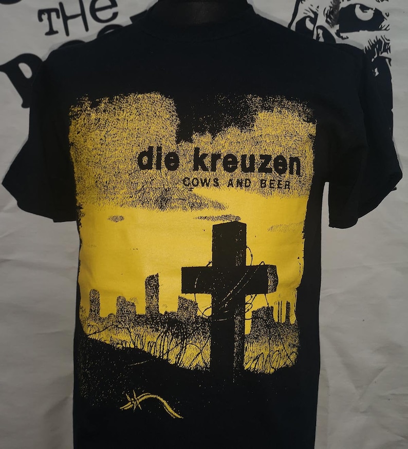 Puede incluir: Camiseta negra con un estampado gr&aacute;fico amarillo de una cruz con alambre de p&uacute;as y el texto "die kreuzen cows and beer".