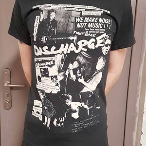 Puede incluir: Camiseta negra con un estampado gr&aacute;fico blanco de la banda de punk rock Discharge. El gr&aacute;fico incluye el nombre de la banda, una foto de los miembros de la banda y el texto "We Make Noise Not Music!!!" y "Fight Back".