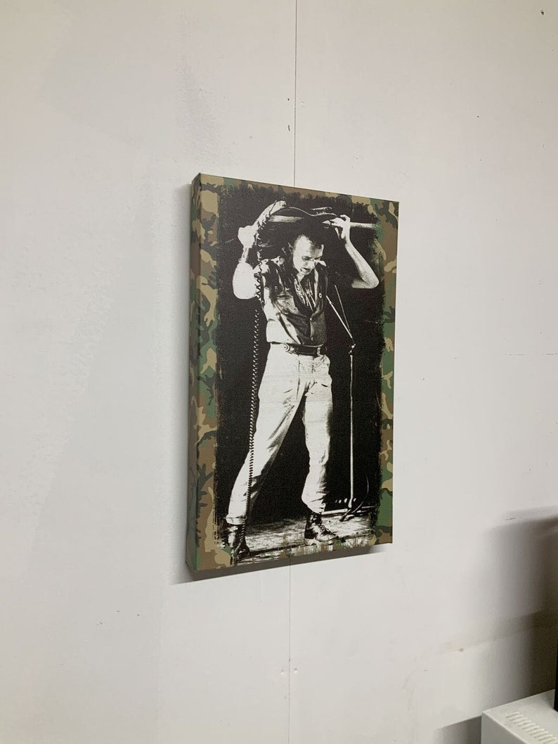 Op de afbeelding: Een zwart-witfoto van een muzikant die een gitaar boven zijn hoofd houdt. De foto is afgedrukt op een canvas met een camouflage-patroonrand.