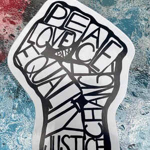 Puede incluir: Una pegatina blanca con forma de puño en alto con contornos negros. El puño está relleno con las palabras "PEACE", "LOVE", "EQUALITY", "CHANGE" y "JUSTICE" en una fuente en negrita sin serifa. La pegatina está sobre una superficie texturizada multicolor.