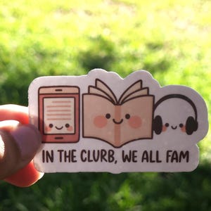 May include: A sticker with a cartoon design of an e-reader, an open book, and headphones, all with smiling faces. The text "IN THE CLURB, WE ALL FAM" is below the images. The sticker is held against a blurred green background.