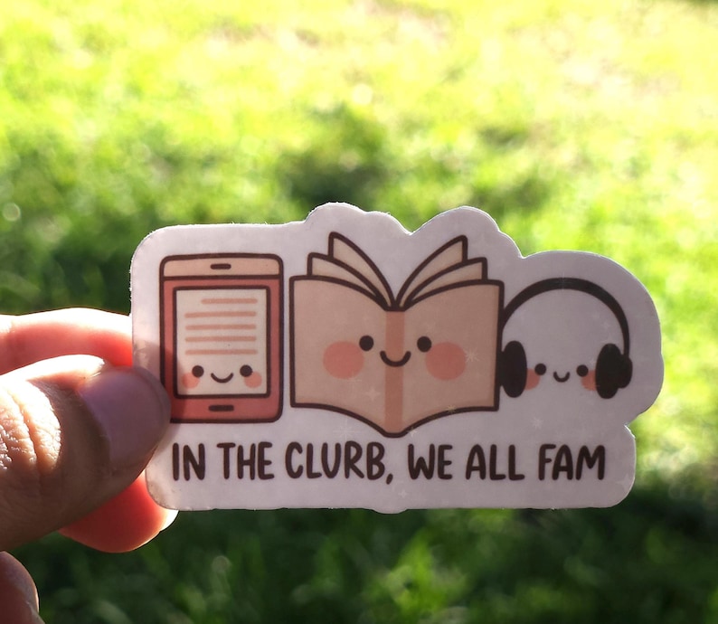 May include: A sticker with a cartoon design of an e-reader, an open book, and headphones, all with smiling faces. The text "IN THE CLURB, WE ALL FAM" is below the images. The sticker is held against a blurred green background.