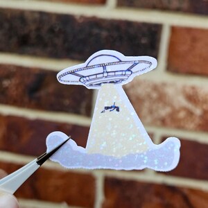 May include: A sticker featuring a white UFO with a blue outline, hovering over a light-colored beam. The beam appears to be lifting a small figure. The sticker has a glittery effect and is set against a brick background.