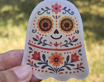 May include: A ghost-shaped sticker featuring a Day of the Dead design. The sticker is cream-coloured with orange, red, and green floral patterns. The eyes are large and orange, and the ghost has a small, smiling mouth.