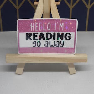 May include: A rectangular sticker with the text "HELLO I'M READING go away" in black and white. The sticker has a pink background with glitter and star accents, and is displayed on a small wooden easel.