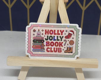 May include: A rectangular sticker with the text "HOLLY JOLLY BOOK CLUB" in red, pink, and green. The sticker features festive illustrations of a candle, books, a gingerbread man, a stocking, a candy cane, a cookie, and boots. The sticker is displayed on a small wooden easel.