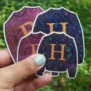 May include: Four stickers in the shape of sweaters are held in front of a green background. The stickers are various shades of purple and blue with gold letters and a sparkly finish. The letters are "H" and "D".