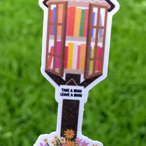May include: A sticker depicting a small library with a brown roof and open windows showing colourful books. The text "TAKE A BOOK LEAVE A BOOK" is below. A bouquet of flowers is at the base.