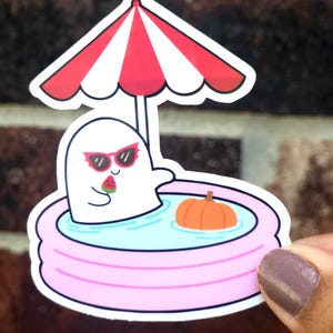 May include: A die-cut sticker featuring a white ghost wearing red cat-eye sunglasses, sitting in a light pink inflatable pool filled with blue water. The ghost holds a slice of watermelon, and a small orange pumpkin floats nearby under a red and white striped umbrella.