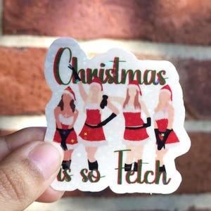 May include: A holographic sticker with the text "Christmas is so Fetch" in red and green. The sticker shows four figures in Santa outfits, wearing red dresses, black gloves, and Santa hats.