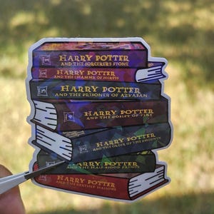 May include: A sticker featuring a stack of seven Harry Potter book covers. Each book has a different color and title, including "Harry Potter and the Sorcerer's Stone" and "Harry Potter and the Deathly Hallows". The sticker is held by tweezers against a blurred green background.