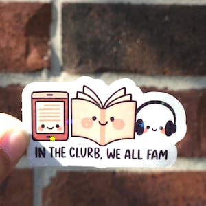May include: A white sticker with a cartoon design of an e-reader, an open book, and headphones, each with a smiling face. The text "IN THE CLURB, WE ALL FAM" is printed below. The sticker is held against a red brick wall.