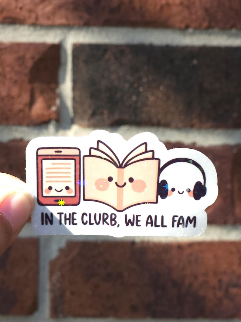 May include: A white sticker with a cartoon design of an e-reader, an open book, and headphones, each with a smiling face. The text "IN THE CLURB, WE ALL FAM" is printed below. The sticker is held against a red brick wall.