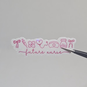 May include: A pink sticker featuring medical symbols: a syringe, plaster, heart, stethoscope, nurse's cap, and pill bottle. The words "future nurse" are written in cursive. The sticker is held by tweezers.
