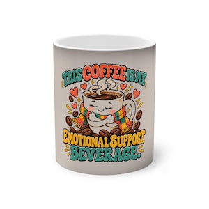 Könnte beinhalten: Ein weißer Keramikbecher mit einer illustrierten Kaffeetasse, die einen bunten gestreiften Schal trägt und sich selbst umarmt. Der Text lautet THIS COFFEE IS MY EMOTIONAL SUPPORT BEVERAGE. Das Design enthält kleine rote Herzen, gelbe Sterne und braune Kaffeebohnen.