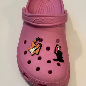 May include: Pink Crocs with two shoe charms featuring a cartoon image of a man in a pink suit and a man in a yellow suit.