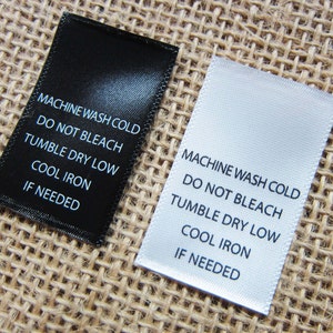 May include: Two white satin fabric care labels with black text. The text on the labels reads "Machine Wash Cold, Do Not Bleach, Tumble Dry Low, Cool Iron, If Needed".