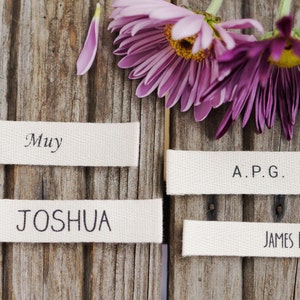 May include: Four white fabric labels with black text on a wooden background. The labels read "Muy", "Joshua", "A.P.G.", and "James Black".