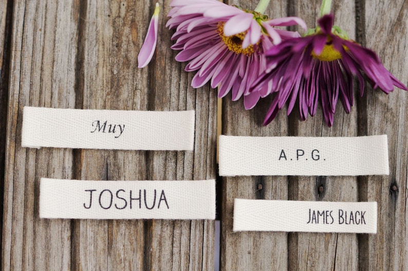 May include: Four white fabric labels with black text on a wooden background. The labels read "Muy", "Joshua", "A.P.G.", and "James Black".