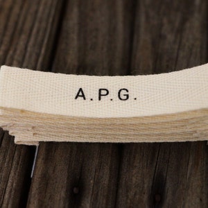 May include: A stack of white fabric labels with the text "A.P.G." printed in black.