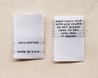 May include: Two white fabric care labels with black text. The label on the left says "100% COTTON" and "MADE IN U.S.A.". The label on the right says "HAND WASH COLD WITH LIKE COLORS", "DO NOT BLEACH", "HANG TO DRY", "COOL IRON", "IF NEEDED".