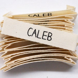 May include: A stack of off-white woven fabric labels with the name "Caleb" printed in black.