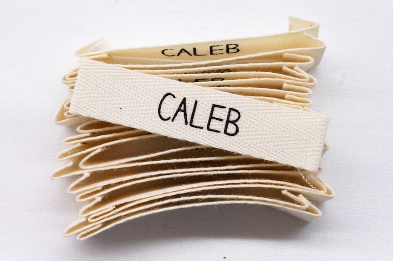 May include: A stack of off-white woven fabric labels with the name "Caleb" printed in black.