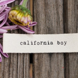 May include: A white fabric label with the text "california boy" printed in black on a wooden background with purple flower petals.