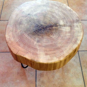 Peut inclure: Une table basse ronde en bois, faite à partir d'une tranche de tronc d'arbre. Le grain naturel du bois est visible, de couleur brun clair. La table a un pied en épingle à cheveux en métal foncé. La table mesure environ 61 cm de diamètre.