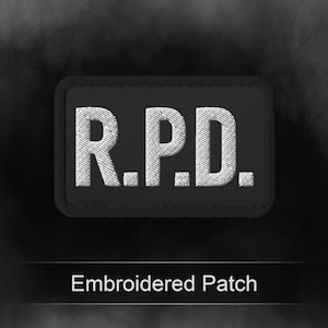 May include: A rectangular black embroidered patch featuring the white stitched text R.P.D. in a bold, sans-serif font. The patch has a thick, stitched border and a textured fabric surface, designed for attachment to clothing or gear items.