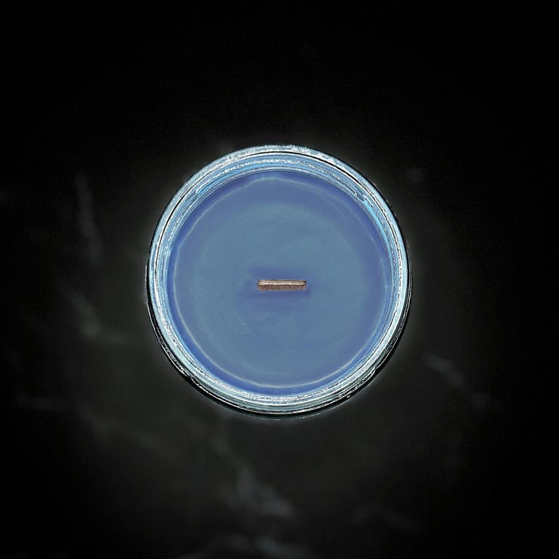 May include: A top-down view of a blue candle in a clear glass jar. The candle has a wooden wick centered in the wax. The jar is set against a dark background.