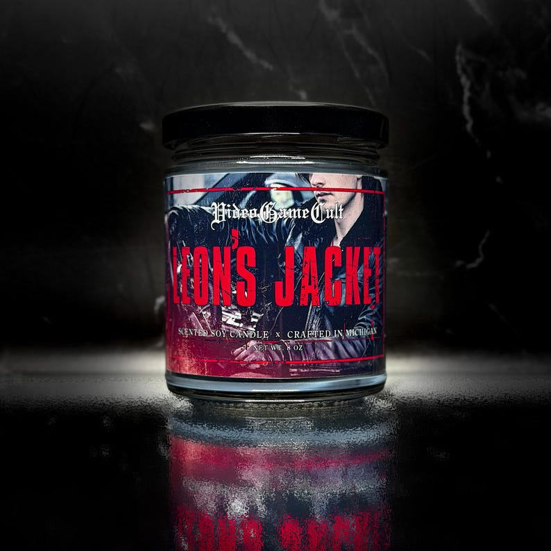 May include: A glass jar candle with a black lid. The label reads "Video Game Cult" and "Leon's Jacket" in red text. The candle has a black and white image of a person wearing a leather jacket. The candle is 8 oz.