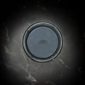May include: Overhead view of a dark gray candle in a glass container. The candle has a single wick centered in the wax. The container has a silver rim and sits on a dark, textured surface.
