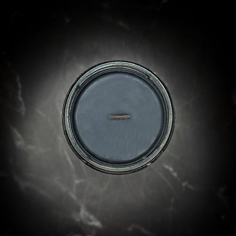 May include: Overhead view of a dark gray candle in a glass container. The candle has a single wick centered in the wax. The container has a silver rim and sits on a dark, textured surface.