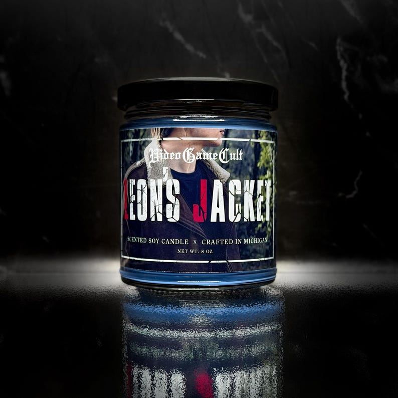 May include: A blue-tinted glass jar with a black lid. The label reads "Video Game Cult" and "Leon's Jacket." The candle is a scented soy candle, net weight 8 oz. The label features a person wearing a jacket.
