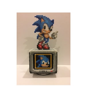 Puede incluir: Una figura de pixel art azul y blanca de Sonic the Hedgehog de pie sobre un televisor de pixel art gris y blanco. La pantalla del televisor muestra una imagen de pixel art de la cabeza de Sonic.