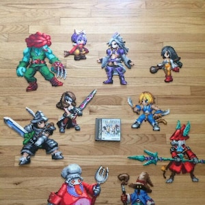 May include: A collection of Final Fantasy characters made with perler beads. The characters include a red-headed warrior, a blue-haired mage, a green-skinned monster, a red-haired woman, a blue-haired woman, a black-haired woman, a blue-haired man, a red-haired man with a spear, and a red-haired woman with a fork. The characters are arranged on a wooden surface. The game cartridge for Final Fantasy VII is also included in the image.