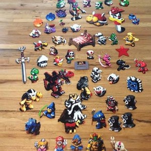 Puede incluir: Una colección de personajes de píxeles coloridos de la serie de videojuegos Super Mario Bros. Los personajes están hechos de pequeñas cuentas y están dispuestos sobre una superficie de madera. Los personajes incluyen a Mario, Luigi, la princesa Peach, Bowser, Toad, Goomba, Koopa Troopa y muchos más.