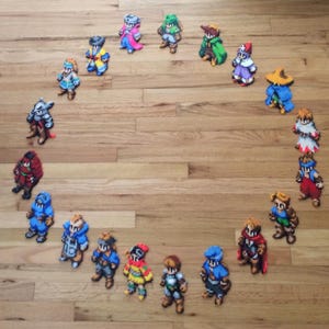 May include: A circle of 16 pixel art characters, each with a unique design and color scheme. The characters are arranged in a circle on a wooden surface.