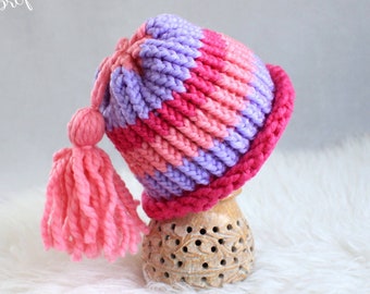 May include: A pink and purple striped knitted baby hat with a pink tassel. The hat is sitting on a wooden stand.