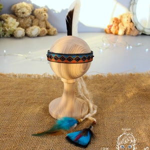 May include: A Native American inspired headband with a black, orange, and blue geometric pattern. The headband is on a wooden stand with a white feather and two blue feathers.
