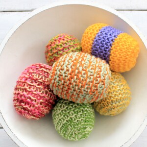 May include: A white bowl filled with six knitted Easter eggs in various colors, including pink, green, yellow, orange, and purple.