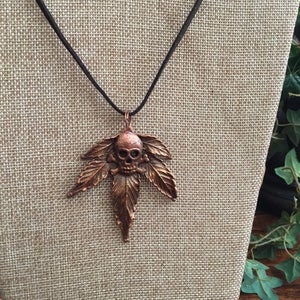 May include: A copper pendant necklace featuring a skull with crossbones in the center of a stylized marijuana leaf.