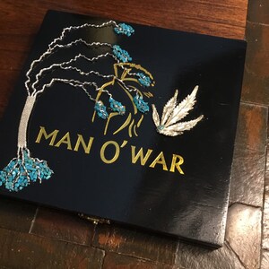 May include: A black wooden box with a gold inscription that reads "MAN O'WAR". The box is decorated with a silver leaf design and turquoise blue stones.