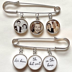 May include: Two large silver safety pins with dangling circular charms. The charms feature black and white photos of people and the text "We have the best seat in the house" in black script. The pins are on a white background.
