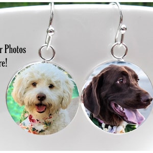 May include: A pair of silver dangle earrings with round photo charms. The charms feature a white dog with a floral collar and a brown dog with a floral collar.