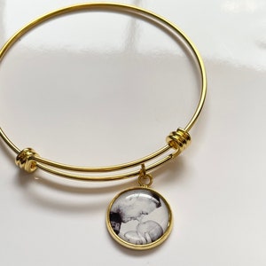 May include: A gold expandable bangle bracelet with a round charm featuring a black and white photo of a couple kissing.